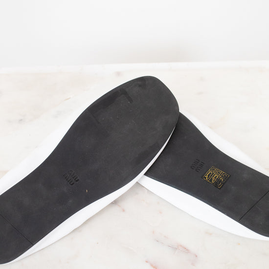 A pair of white slip-on shoes shown from the bottom, revealing black outsoles with a small gold logo on a light marble surface.