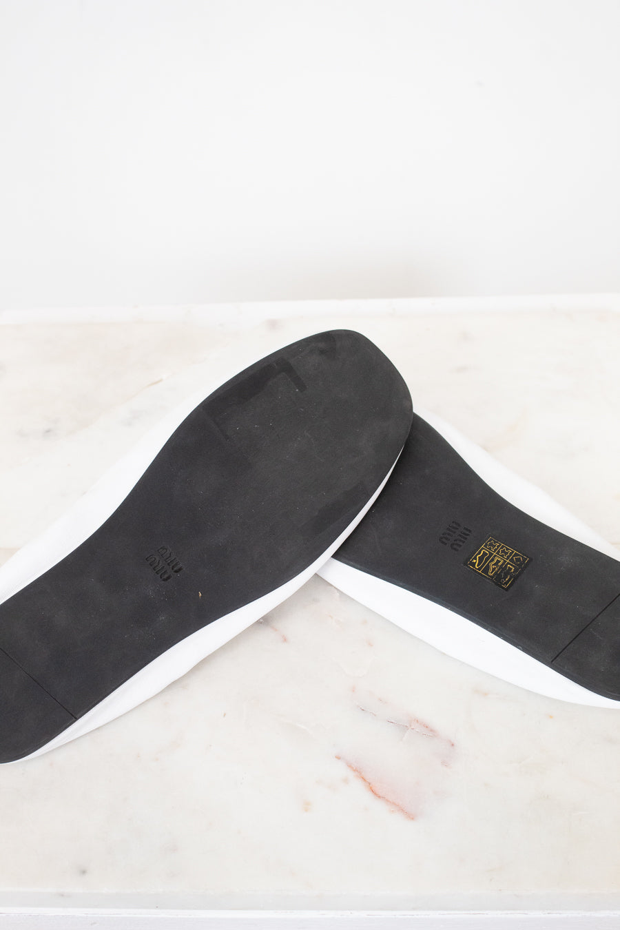 A pair of white slip-on shoes shown from the bottom, revealing black outsoles with a small gold logo on a light marble surface.