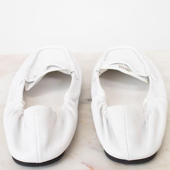 A pair of white leather ballet flats with gathered detailing and a small gold brand plaque, resting on a light marble surface against a white background.