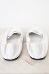 A pair of white leather ballet flats with gathered detailing and a small gold brand plaque, resting on a light marble surface against a white background.