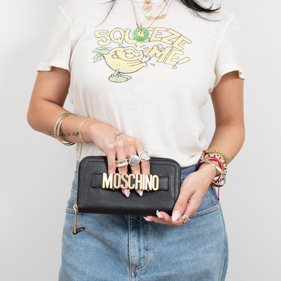 Person wearing a white t-shirt with a graphic design, holding a black wallet with 'Moschino' branding.
