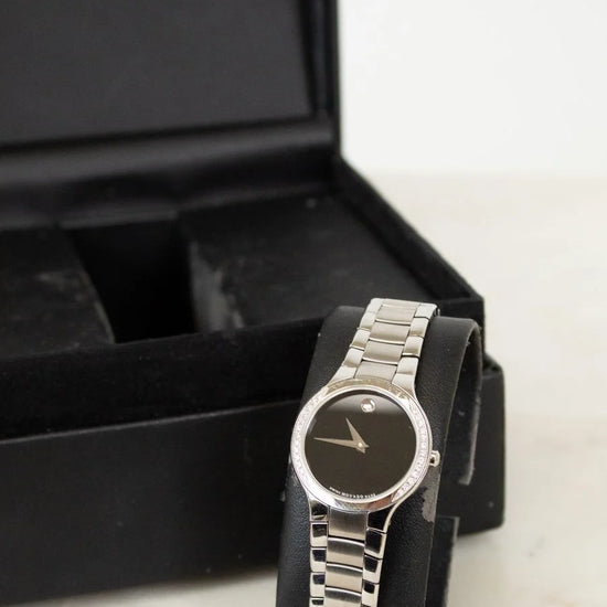 Silver watch with black face on a marble surface next to a black box with 'Movado' branding.