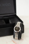 Silver watch with black face on a marble surface next to a black box with 'Movado' branding.