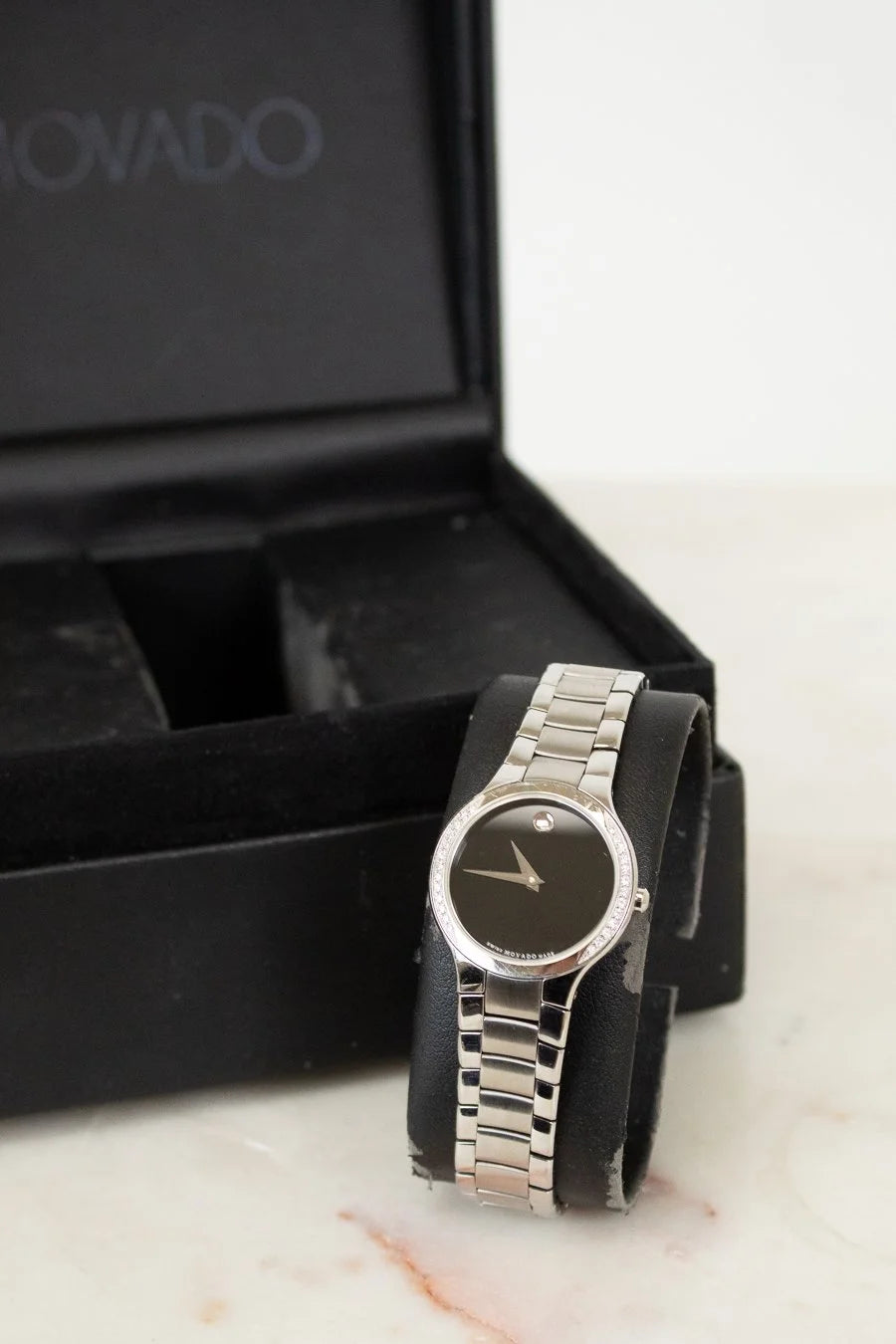 Silver watch with black face on a marble surface next to a black box with 'Movado' branding.