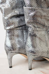 Close-up of metallic silver snakeskin-patterned heeled boots with ruched shafts, shown on a light marble surface.