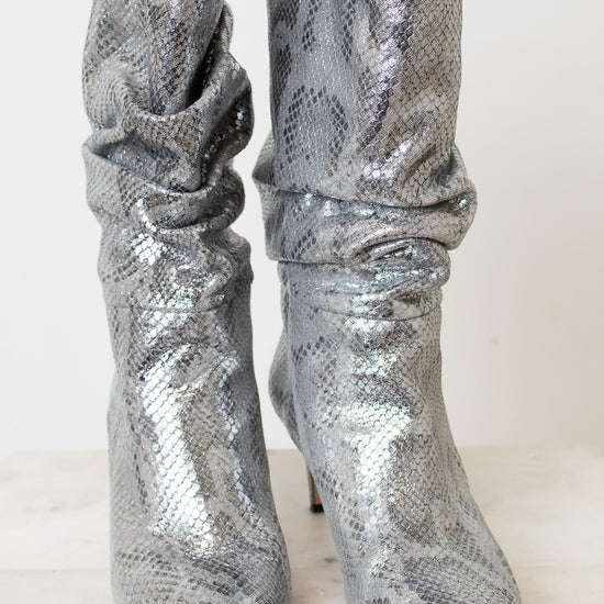 A pair of metallic silver snakeskin ankle boots with a slouchy draped design and pointed toes, set against a plain off-white background.