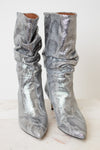 A pair of metallic silver snakeskin ankle boots with a slouchy draped design and pointed toes, set against a plain off-white background.