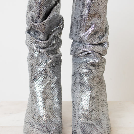 A pair of metallic silver snakeskin boots with a slouchy draped design and pointed toes, set against a plain off-white background.
