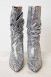 A pair of metallic silver snakeskin boots with a slouchy draped design and pointed toes, set against a plain off-white background.