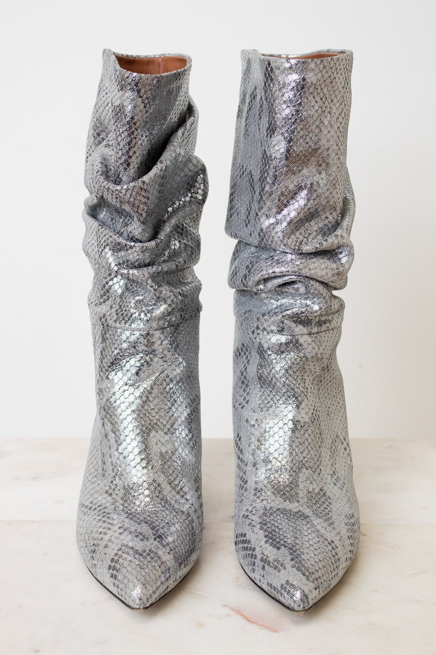 A pair of metallic silver snakeskin boots with a slouchy draped design and pointed toes, set against a plain off-white background.