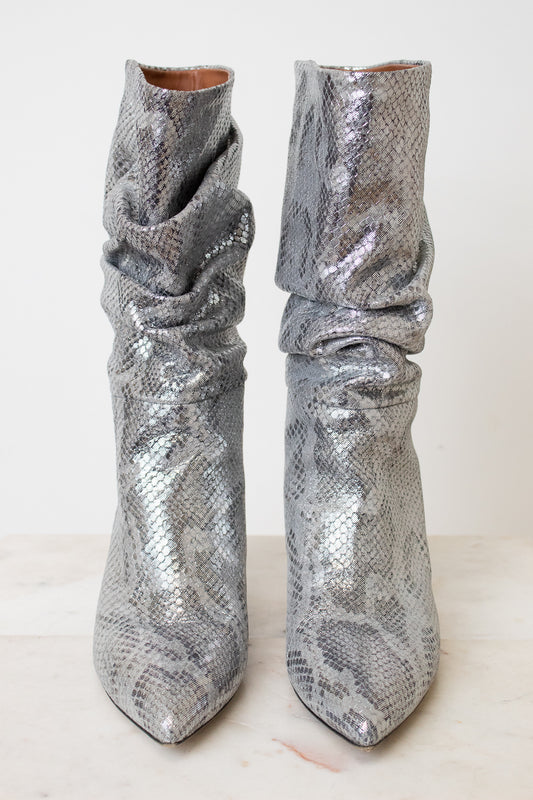 A pair of metallic silver snakeskin boots with a slouchy draped design and pointed toes, set against a plain off-white background.