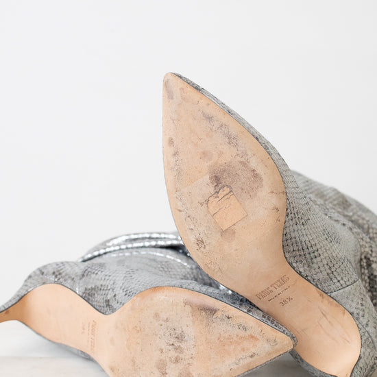 A pair of women's pointed-toe heeled shoes in a metallic silver snakeskin pattern, shown from the sole side on a light marble surface against a white background.
