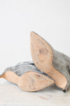 A pair of women's pointed-toe heeled shoes in a metallic silver snakeskin pattern, shown from the sole side on a light marble surface against a white background.