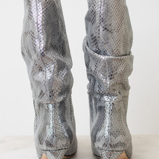 A pair of metallic silver snakeskin slouchy boots with high stiletto heels, shown from the back on a light marble surface against a white background.