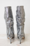 A pair of metallic silver snakeskin slouchy boots with high stiletto heels, shown from the back on a light marble surface against a white background.