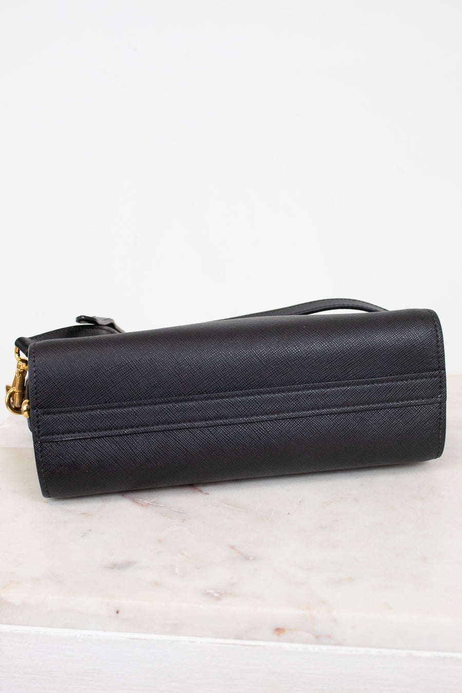 Bottom view of a black textured leather handbag  with gold-tone hardware, resting on a white marble surface against a white background.
