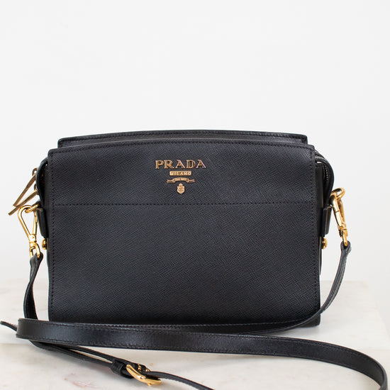A black Prada leather crossbody bag with gold-tone hardware and a gold logo on the front, resting on a light marble surface against a white background.