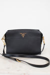 A black Prada leather crossbody bag with gold-tone hardware and a gold logo on the front, resting on a light marble surface against a white background.