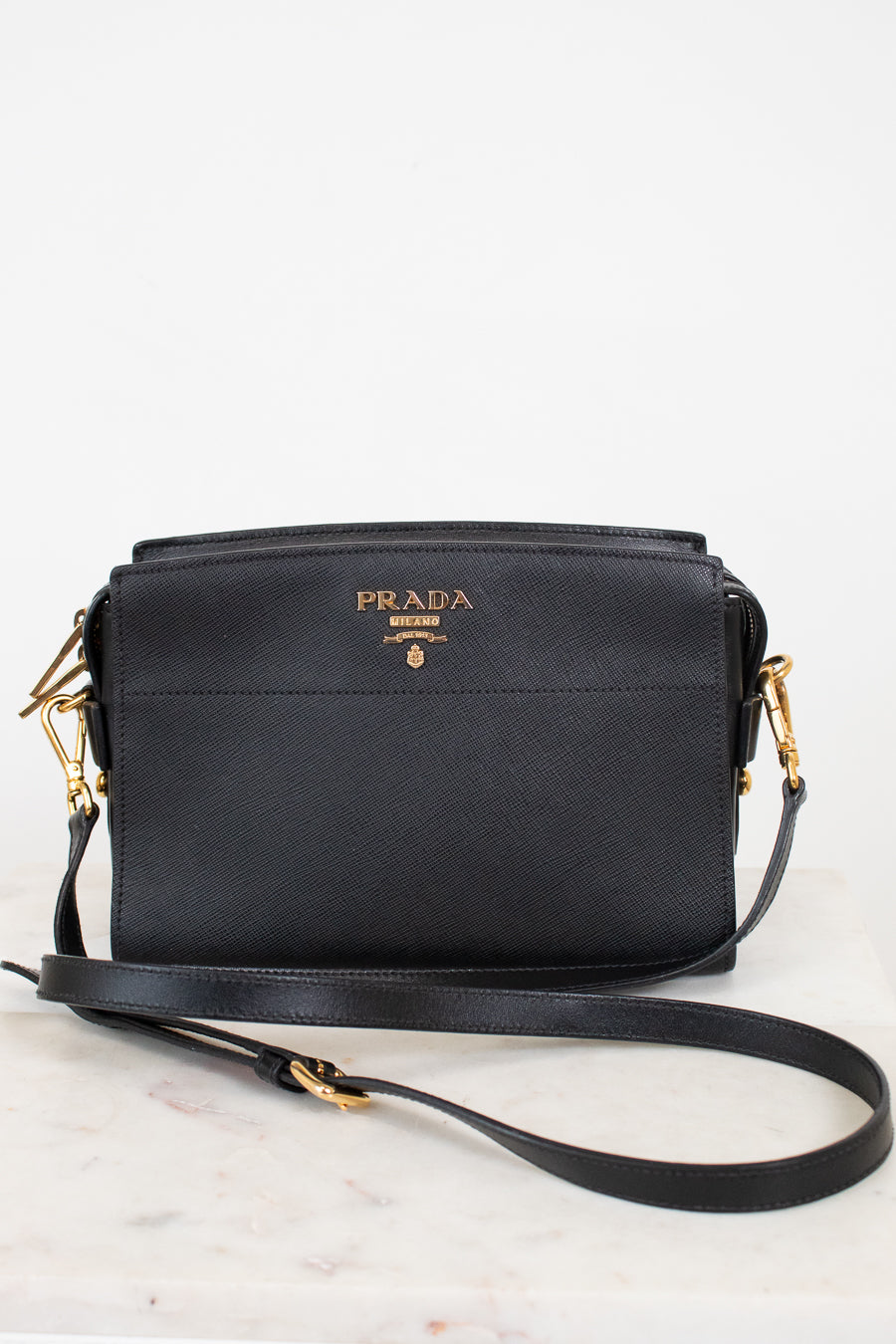 A black Prada leather crossbody bag with gold-tone hardware and a gold logo on the front, resting on a light marble surface against a white background.