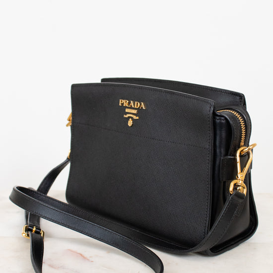 A black Prada crossbody bag with gold-tone hardware and logo, resting on a white marble surface against a white background.