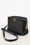 A black Prada crossbody bag with gold-tone hardware and logo, resting on a white marble surface against a white background.