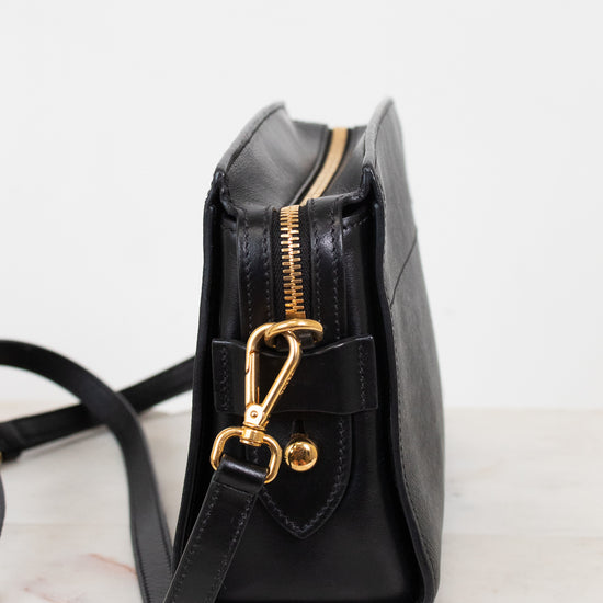 A black leather crossbody bag with gold-tone hardware, shown from a side profile view on a light marble surface against a white background.