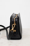 A black leather crossbody bag with gold-tone hardware, shown from a side profile view on a light marble surface against a white background.