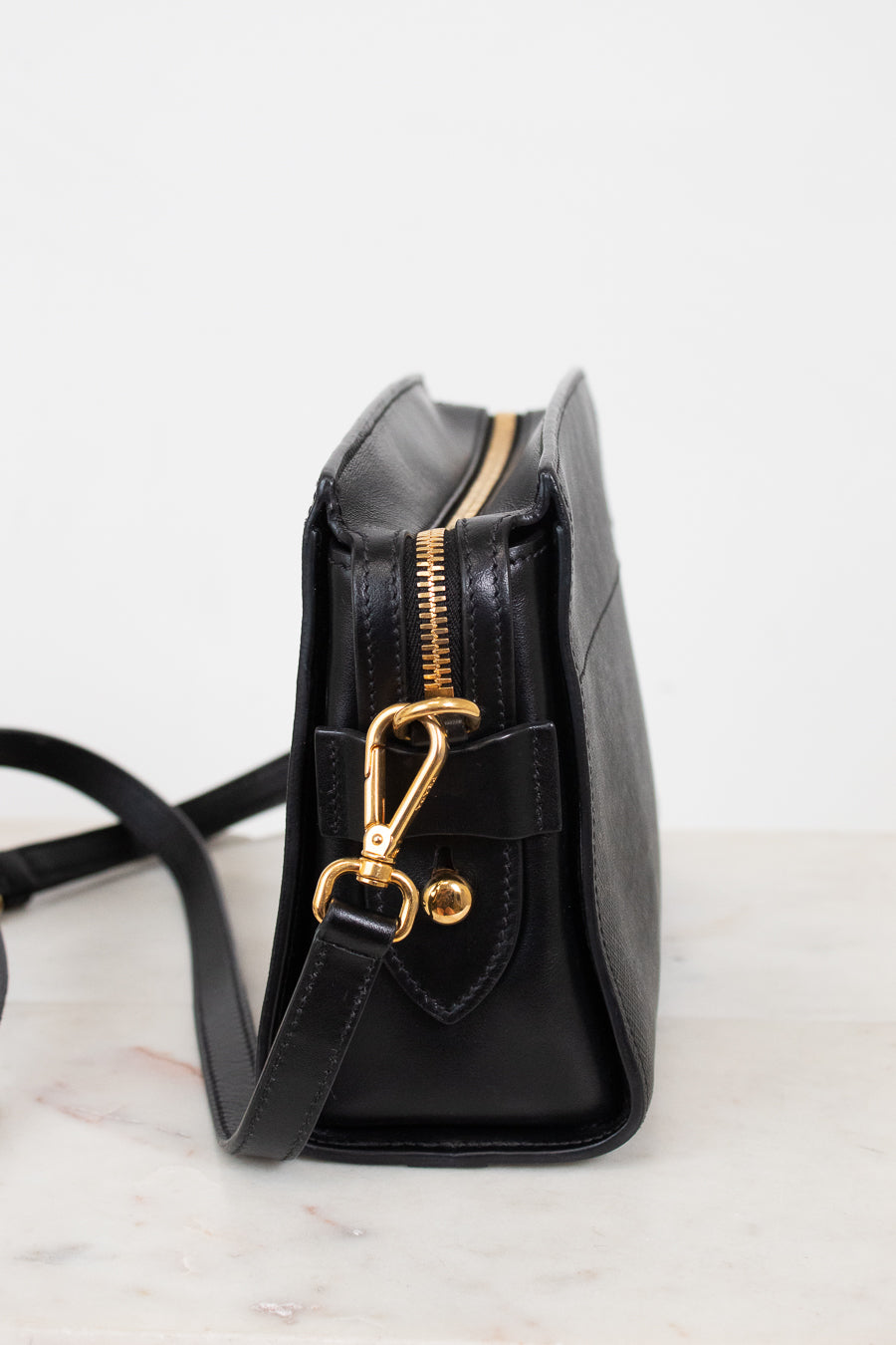 A black leather crossbody bag with gold-tone hardware, shown from a side profile view on a light marble surface against a white background.