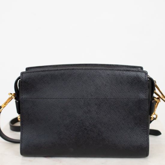 Back view of a black textured leather crossbody bag with gold-tone hardware, resting on a white marble surface against a white background.