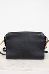 Back view of a black textured leather crossbody bag with gold-tone hardware, resting on a white marble surface against a white background.