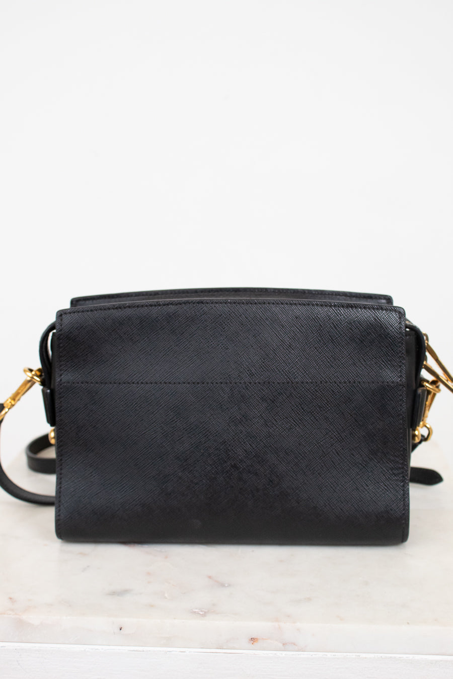 Back view of a black textured leather crossbody bag with gold-tone hardware, resting on a white marble surface against a white background.