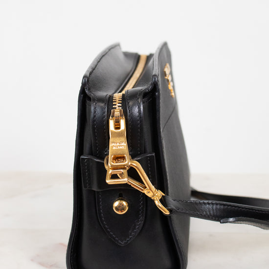 A close-up side view of a black leather handbag with gold-tone hardware, including a zipper pull, clasp, and a small gold logo on the front, set against a clean white background.