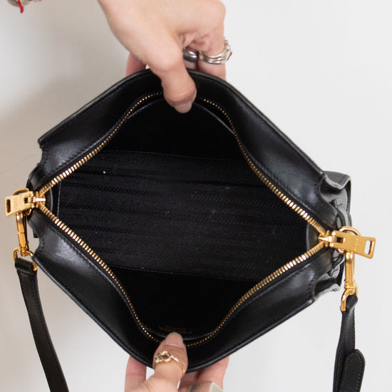 A black leather handbag with gold-tone hardware is held open by two hands against a plain white background.