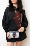 A woman wearing a black fuzzy knit sweater with a large dark red floral pattern, black shorts, and holding a black and silver Prada shoulder bag with a gold chain strap.