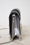A close-up side view of a metallic silver and black leather clutch bag with a silver chain strap, resting on a light marble surface against a white background.