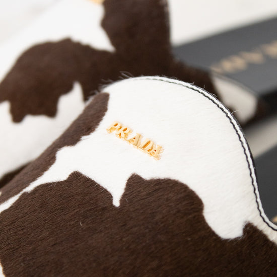 Close-up of a Prada slipper in brown and white cowhide with gold-tone 'PRADA' branding on the side, set against a neutral light background.