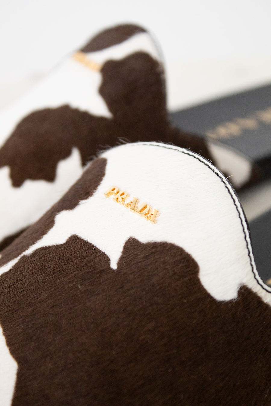 Close-up of a Prada slipper in brown and white cowhide with gold-tone 'PRADA' branding on the side, set against a neutral light background.