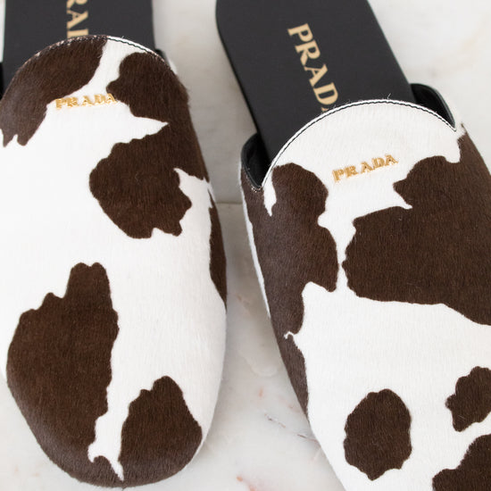 A pair of Prada slip-on mule slippers with a brown and white cow print pattern, displayed on a light marble surface.