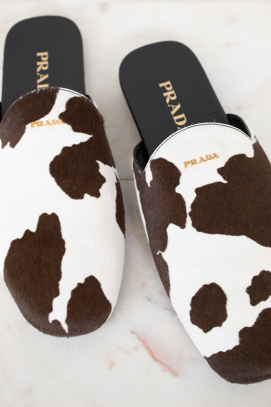 A pair of Prada slip-on mule slippers with a brown and white cow print pattern, displayed on a light marble surface.
