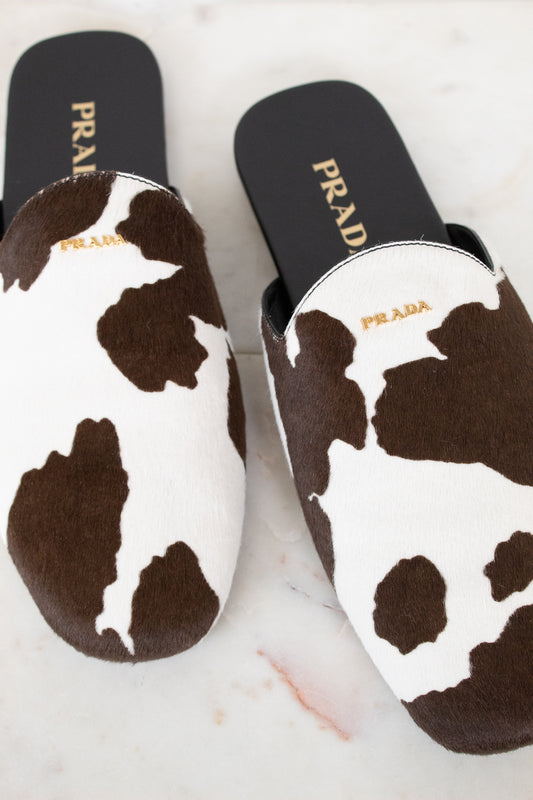 A pair of Prada slip-on mule slippers with a brown and white cow print pattern, displayed on a light marble surface.