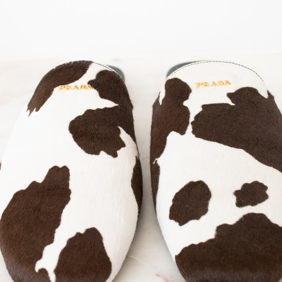 A pair of Prada cow-print slippers in brown and white faux fur, shown from a top-down view on a light marble surface.