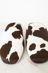 A pair of Prada cow-print slippers in brown and white faux fur, shown from a top-down view on a light marble surface.