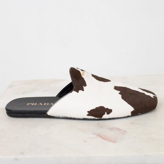 A Prada slip-on mule sandal featuring a brown and white cow-print pattern, resting on a white marble surface against a white background.