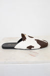 A Prada slip-on mule sandal featuring a brown and white cow-print pattern, resting on a white marble surface against a white background.