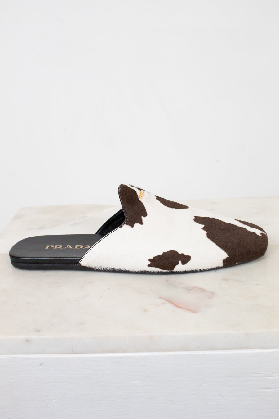 A Prada slip-on mule sandal featuring a brown and white cow-print pattern, resting on a white marble surface against a white background.