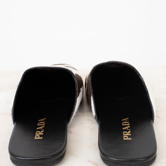 A pair of Prada slide sandals with a black leather base, brown and white patterned side panels, and gold 'PRADA' branding on the insoles, displayed on a light marble surface against a white background.