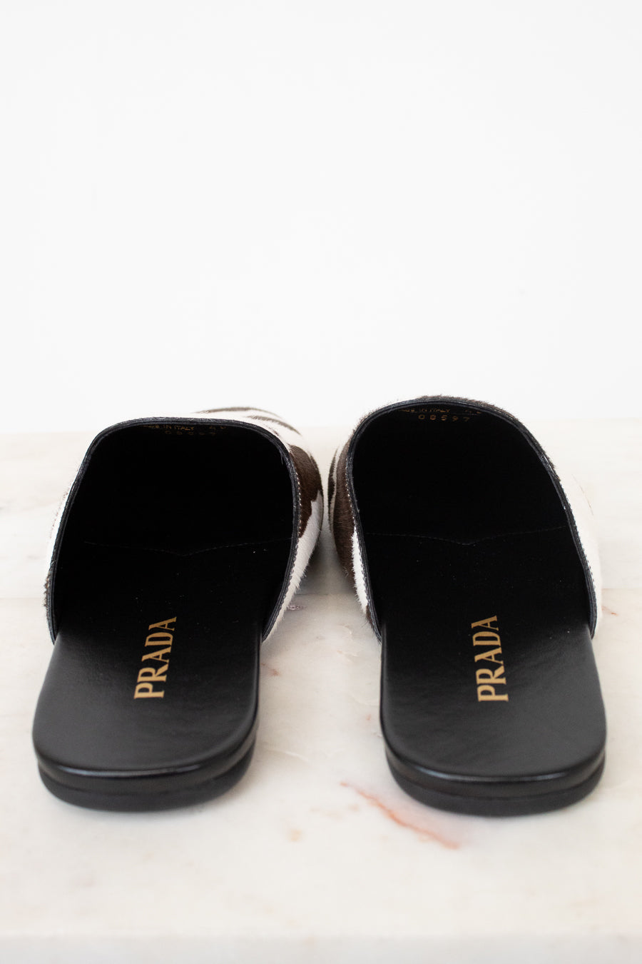 A pair of Prada slide sandals with a black leather base, brown and white patterned side panels, and gold 'PRADA' branding on the insoles, displayed on a light marble surface against a white background.