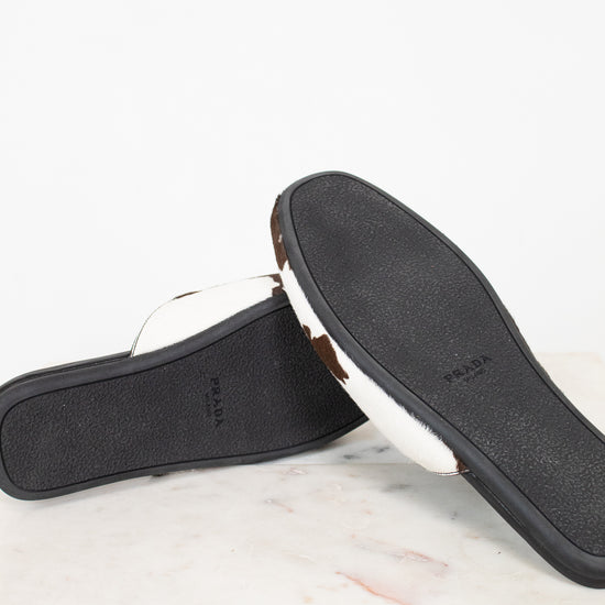A pair of Prada slide sandals shown from the bottom, displaying the black textured sole with the Prada logo and a white leather strap, resting on a white marble surface against a white background.