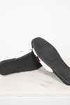 A pair of Prada slide sandals shown from the bottom, displaying the black textured sole with the Prada logo and a white leather strap, resting on a white marble surface against a white background.
