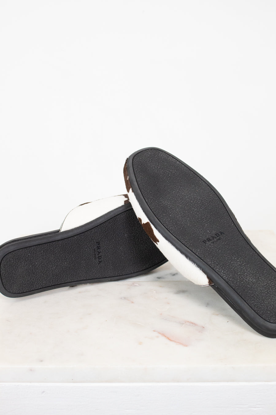 A pair of Prada slide sandals shown from the bottom, displaying the black textured sole with the Prada logo and a white leather strap, resting on a white marble surface against a white background.